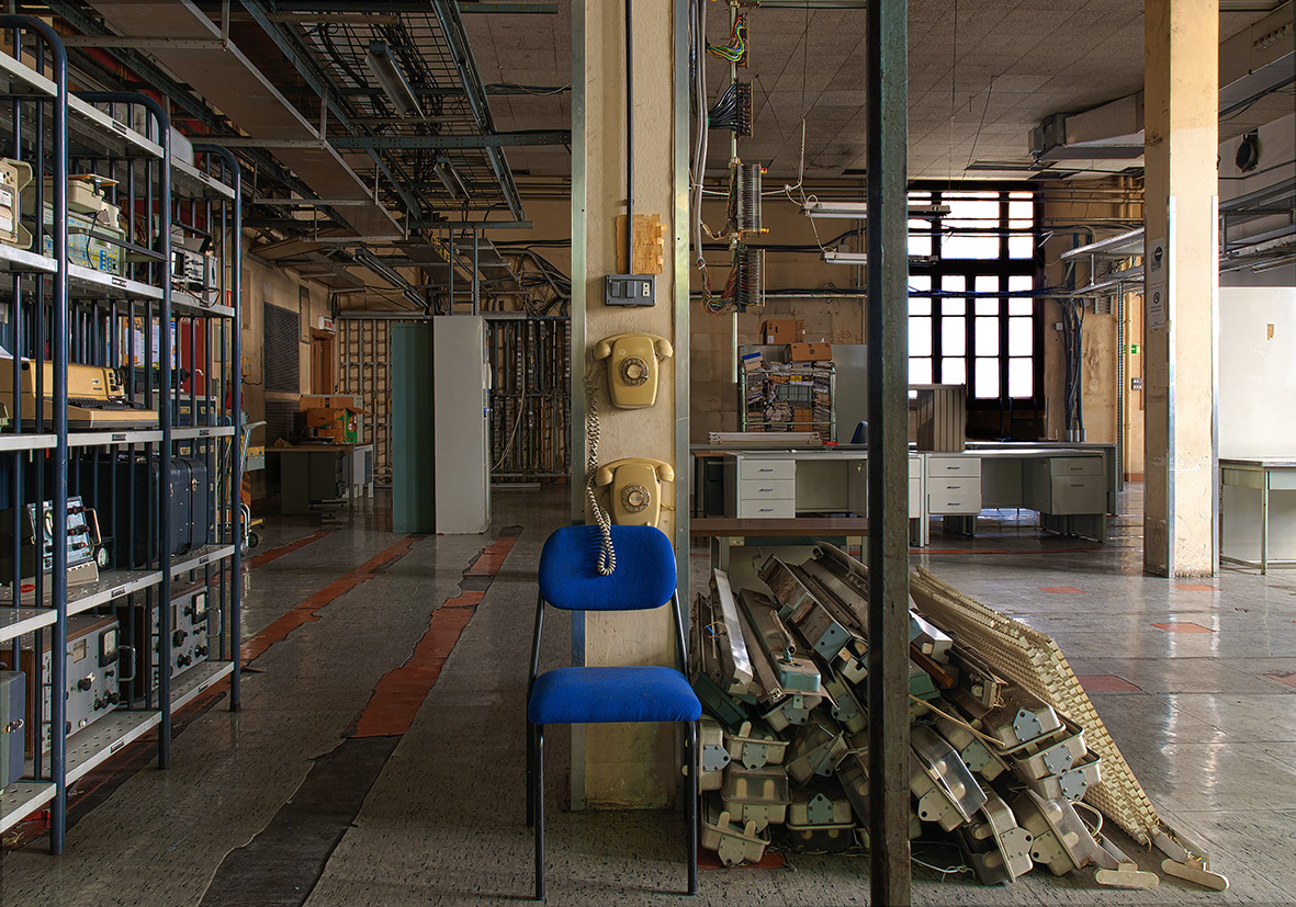 Sala industrial con estanterías, una silla azul, y materiales apilados en el suelo.