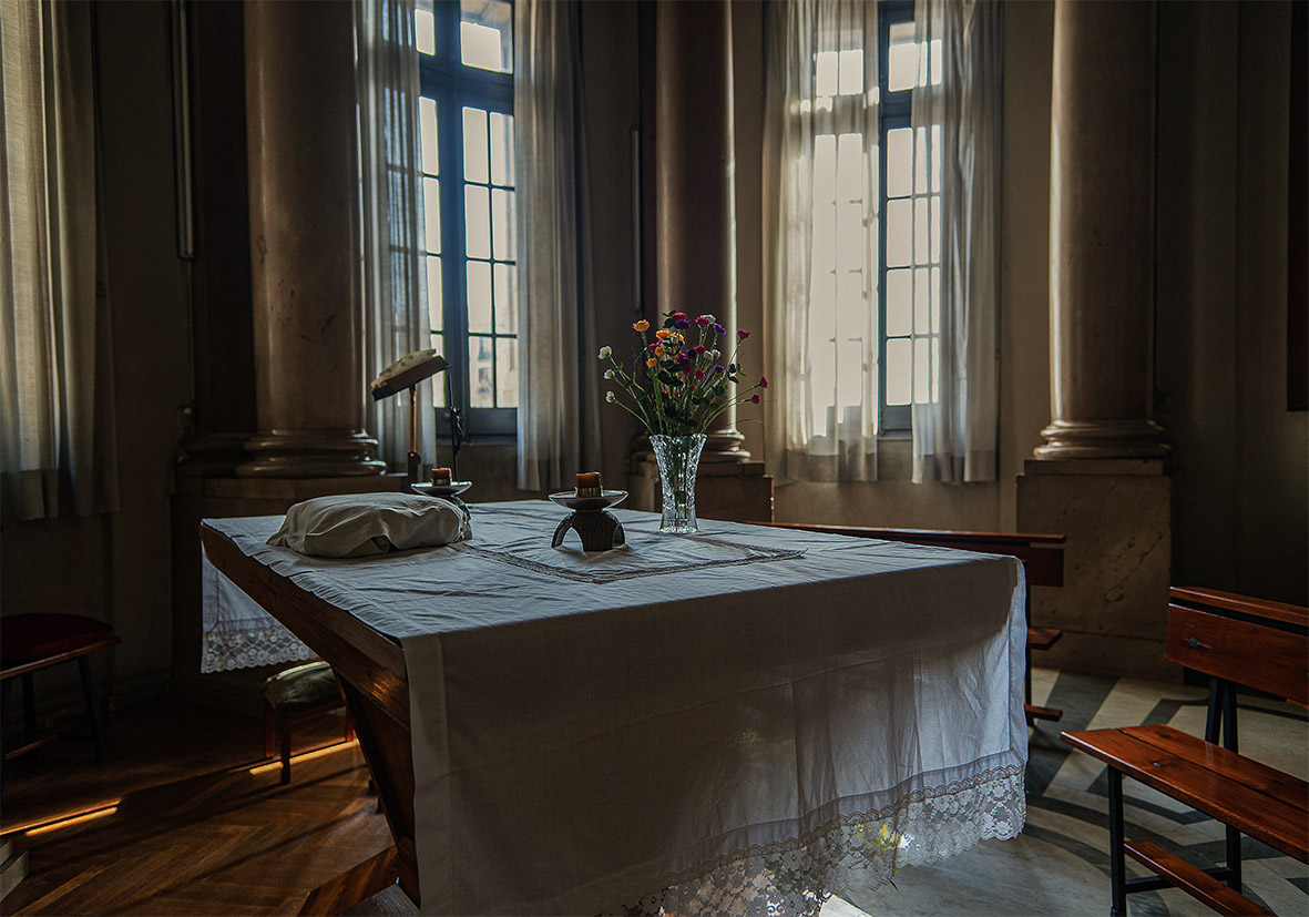 Mesa con mantel blanco y jarrón de flores en una habitación iluminada por ventanas.