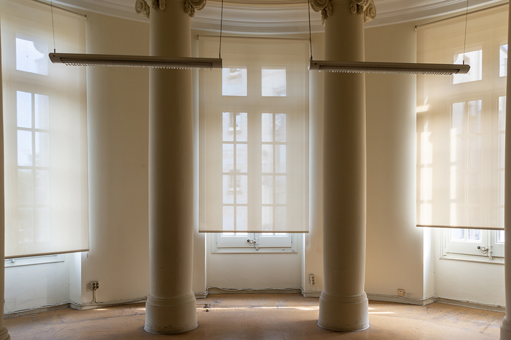 Interior de una habitación con columnas y ventanas con persianas en un entorno luminoso.